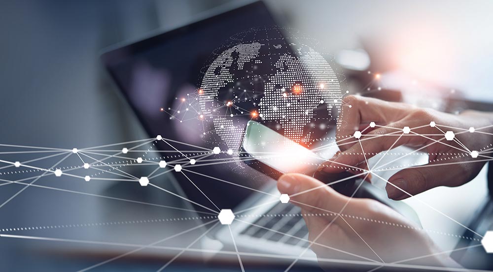 Hände halten und bedienen ein Smartphone, im Hintergrund eine Weltkugel vor einem Laptop, Foto: iStock/TippaPat